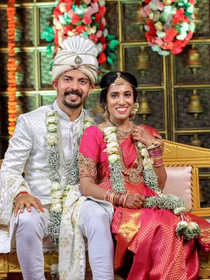 A smiling couple poses for a portrait on their wedding couch, against a backdrop of traditional temple bells.