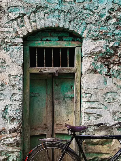 A weathered green door is set within a rough, turquoise-painted stone wall. A bicycle parked in front adds a touch of daily life to this scene rich with texture and history.