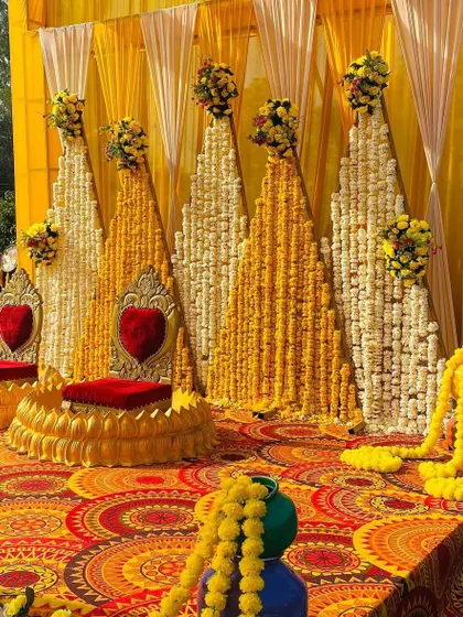 A beautiful Haldi stage with a backdrop of marigold and tuberose strings arranged in a mountain-like pattern. The ornate golden seats with red cushions add a royal touch to the ceremony.
