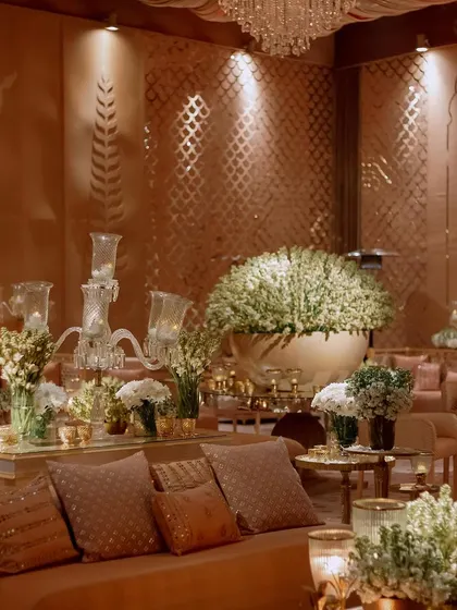 A luxurious lounge area with plush sofas and a mirrored coffee table. The space is filled with large brass pots of white flowers and elegant glass candelabras.