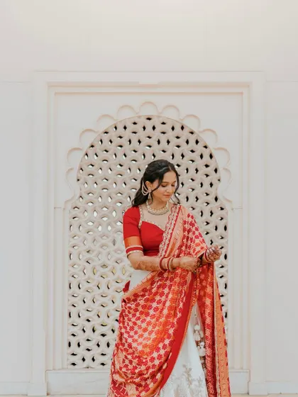 The bride, Kriti, looking radiant in her red and white lehenga. This full-length portrait captures her adjusting her dupatta in a moment of quiet grace.
