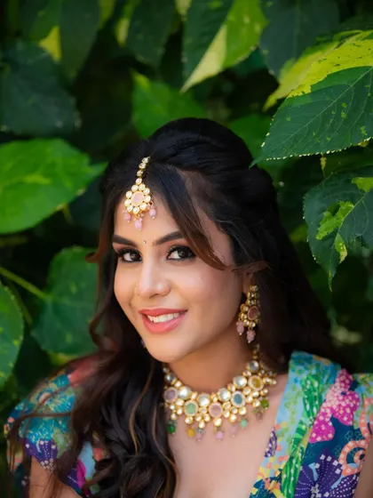 A close-up portrait from a Mehendi look shoot. The focus is on the model's expressive face, festive makeup, and the colorful jewelry that complements her multi-colored outfit.