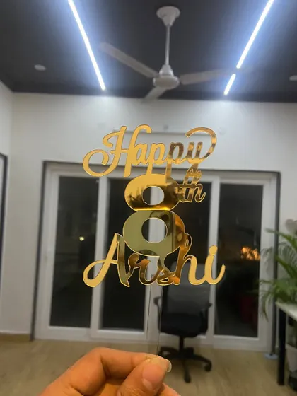 The "Happy 8th Arshi" topper held against an indoor background, showing how its reflective surface shines even in regular lighting.