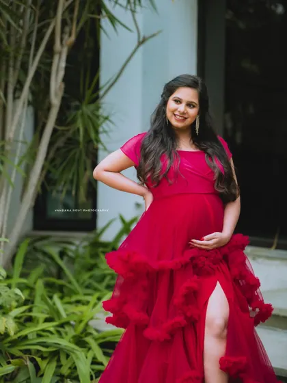 A confident and radiant mother-to-be poses in a stylish red ruffled gown, showcasing her beautiful baby bump in a garden.