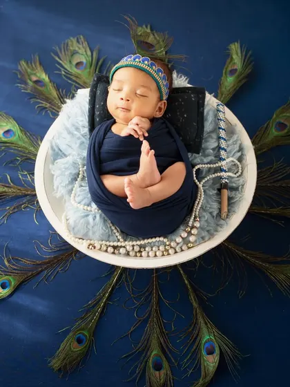 A stunning overhead shot of a newborn Krishna, surrounded by a radial display of peacock feathers on a rich blue background.