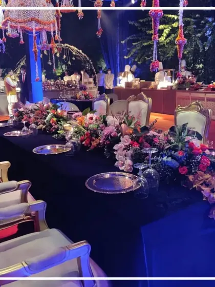 We design dining experiences that match the mood of your celebration. This image captures a luxurious sit-down dinner setting for a sangeet night, featuring a long table dressed in black velvet, accented with silver charger plates and a magnificent floral runner.
