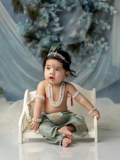 A sweet baby boy dressed as Krishna, sitting on a miniature white bench against a dreamy, icy-blue backdrop. The peacock feathers add a perfect thematic touch.