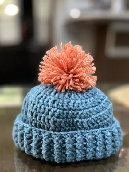 A close-up of one of my handmade baby beanies. The bright orange pom-pom adds such a fun pop of color to the soft blue yarn. It's perfect for keeping a little head warm and stylish.
