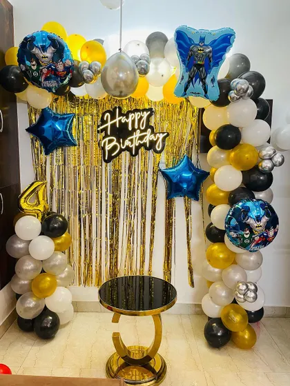 A Batman-themed 4th birthday party setup. This decoration uses a gold shimmer curtain, a black, gold, and white balloon arch, and multiple Batman foil balloons for a heroic celebration.