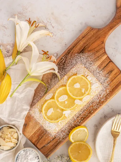 A lemon and almond loaf cake on a wooden board. The bright, airy styling gives this shot a fresh and wholesome feel, perfect for a bakery specializing in tea cakes.