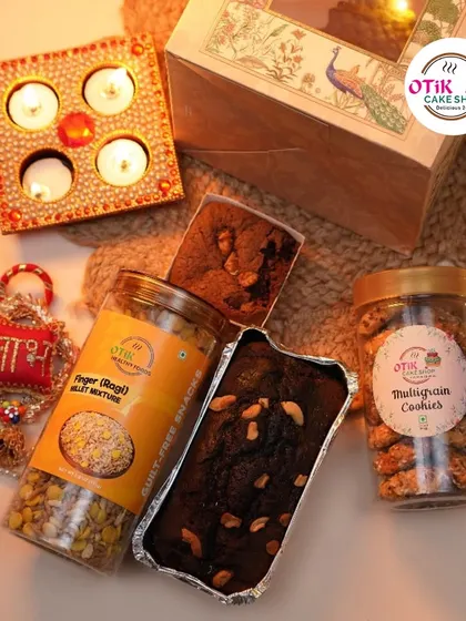 A festive flat-lay showing a Diwali hamper with diyas and rakhis. This box includes a finger millet mixture, multigrain cookies, and a rich dry cake.