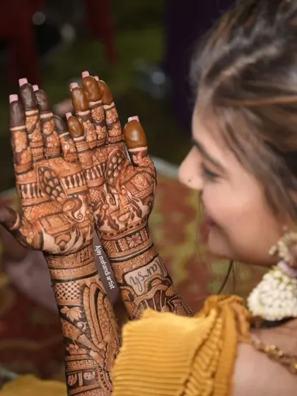 A candid shot of a bride during her mehndi ceremony, her hands held up in a prayer-like pose, showing the detailed design.