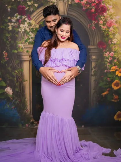 A classic couple's pose, forming a heart with their hands on the baby bump. The floral archway backdrop adds a layer of romance and magic.