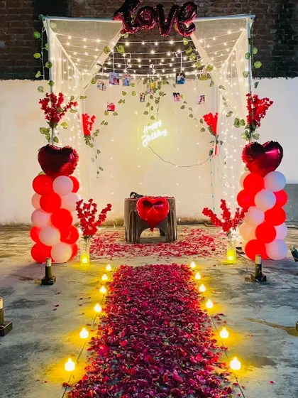 An intimate and romantic outdoor cabana setup, perfect for a birthday or anniversary surprise. The decor includes a candle-lit rose petal pathway, balloon pillars, and a canopy decorated with fairy lights and a 'Happy Birthday' neon sign.