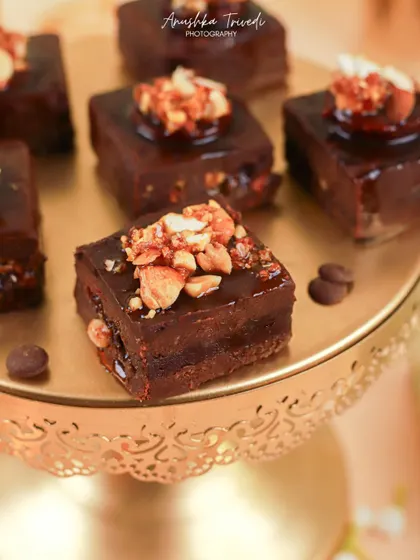 A detailed close-up of rich, glossy chocolate brownies topped with crunchy nuts, presented on a golden cake stand.