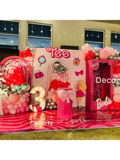 A vibrant and fun Barbie-themed birthday party stage. The setup includes a life-size Barbie doll box for photos, pink and red balloon garlands, and a light-up number '3'.