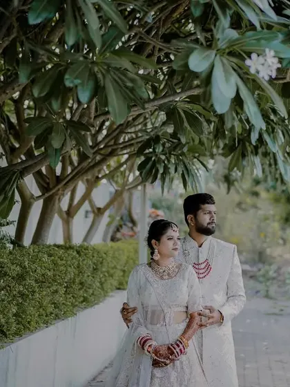 A stylish portrait of a couple in elegant white and cream outfits, walking along a path lined with greenery.
