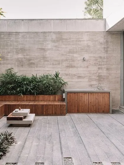 This outdoor patio features built-in wooden seating against a raw concrete wall. The design integrates planters into the seating structure, creating a functional and stylish space that blends architecture with nature.