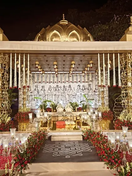 A frontal view of the magnificent brass mandap, showcasing its intricate design and symmetry. The backdrop features a hand-drawn illustration of deities, while the foreground is filled with red roses and traditional brass lamps.
