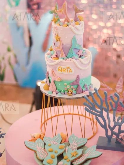 The cake and cookie display for a mermaid-themed party. The cake is a two-tiered masterpiece with mermaid tails, and the cookies are shaped like mermaid fins, all in a lovely pastel palette.