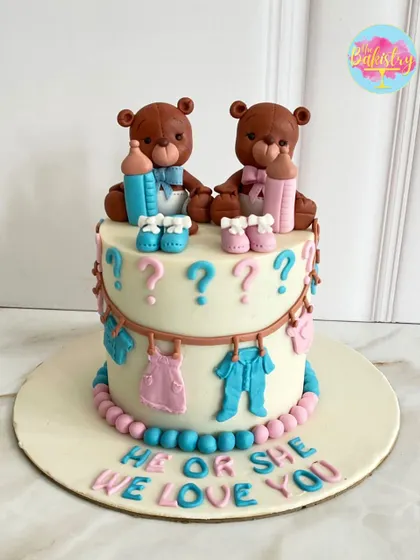He or she? We love you! This adorable gender reveal cake features two teddy bears, one with a pink bow and one with a blue bow, surrounded by tiny baby clothes.