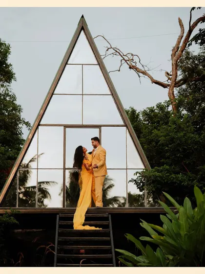 A stunning architectural shot of a couple in yellow outfits posing in front of a modern A-frame glass house.