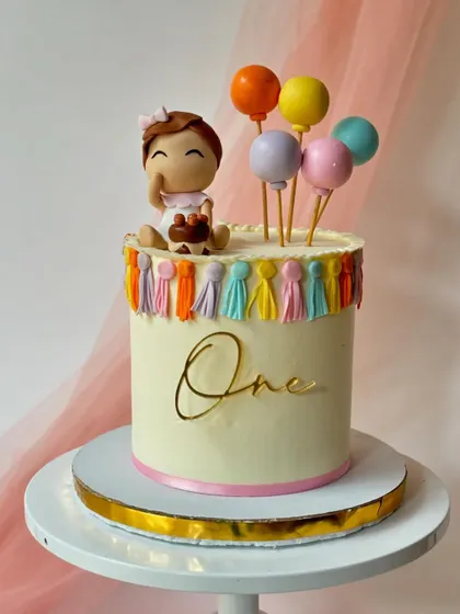 A festive first birthday cake with a girl topper holding her own mini cake. The design is decorated with colourful tassels and balloons.