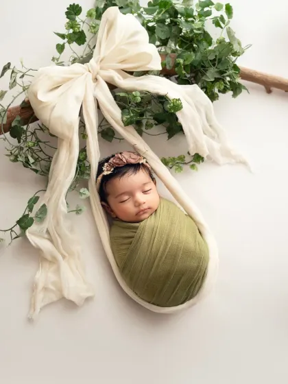 A classic newborn pose, safely suspended in a sling. The simple white background and touch of greenery create a natural, organic feel for this baby photoshoot.