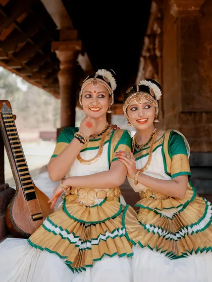A portrait of two dancers posing together, with one holding an instrument. The image celebrates the art of Mohiniyattam and the connection between the performers.