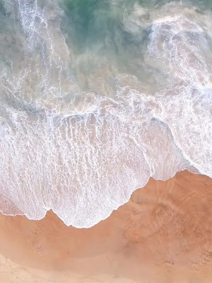 A top-down aerial photograph of the Goa coastline, showing the beautiful contrast between the turquoise water, white seafoam, and the warm-toned sandy beach.
