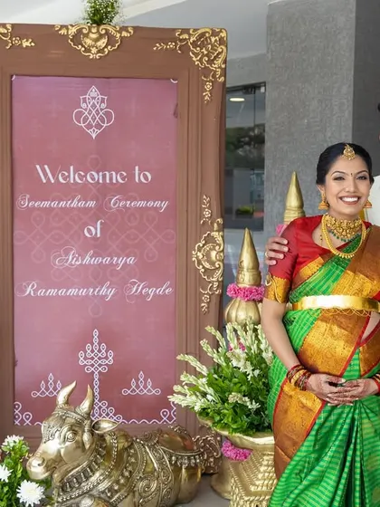 The couple welcomes guests to the Seemantham ceremony with a custom-designed sign, flanked by traditional brass and floral decorations.