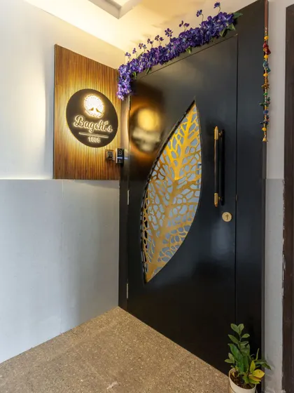 An elegant and artistic entryway, featuring a black door with a golden leaf-patterned jali cutout and a backlit wooden nameplate.