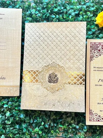 A classic invitation in beige and gold, featuring a textured finish and a central Ganesha emblem. The insert has a beautiful maroon border with traditional patterns.