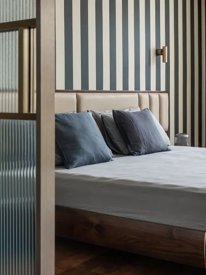 This bedroom is a study in refined comfort, featuring a bold striped wallpaper that adds graphic interest without overwhelming the space. A fluted glass partition provides a subtle separation, enhancing privacy while allowing light to filter through, creating a serene and layered retreat.