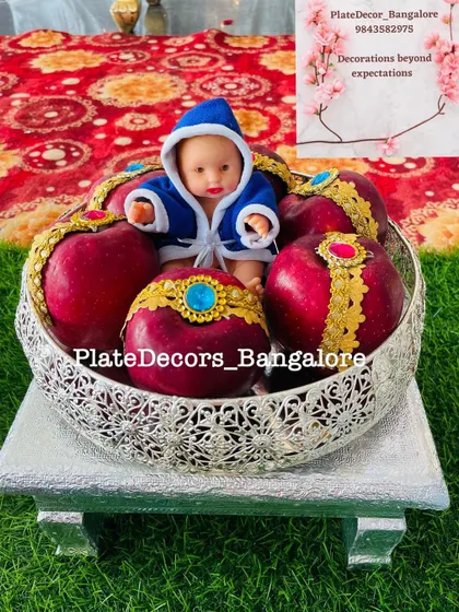 This silver bowl contains red apples adorned with golden lace and blue kundan stones, with a baby doll in a blue robe as the centerpiece. A perfect gift for a baby boy's arrival ceremony.