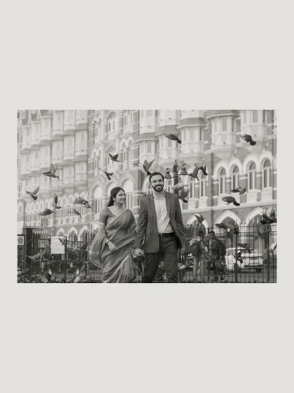 A beautiful black and white shot of the couple walking past the Taj Mahal Palace hotel, with pigeons taking flight around them, creating a classic and romantic scene.