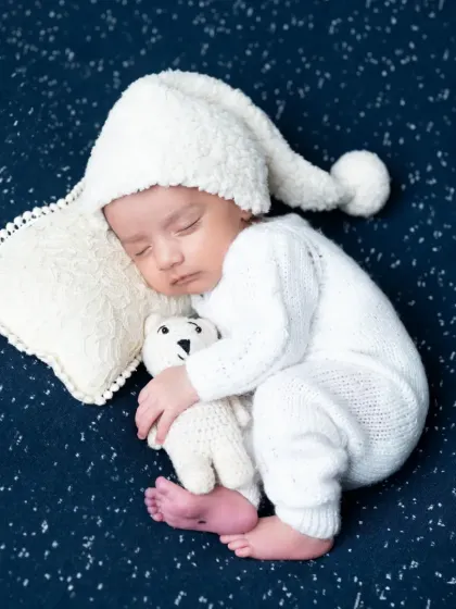 A full-body shot of the baby boy in a different white hat, hugging his teddy bear.