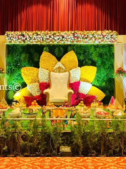A grand stage setup featuring a large lotus backdrop and a full ceremonial plate arrangement. The potted plants in the foreground add a touch of natural greenery.
