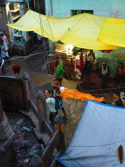 An overhead view shows a body prepared for cremation lying in a narrow alleyway under makeshift tarps. This perspective provides a different, more documentary context to the pre-cremation rituals.