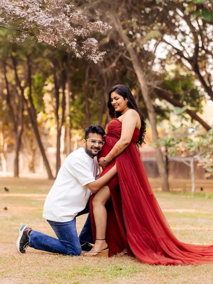 A candid and joyful moment during an outdoor maternity shoot. The partner hugs the baby bump from a kneeling position, both sharing a smile under a blossoming tree.