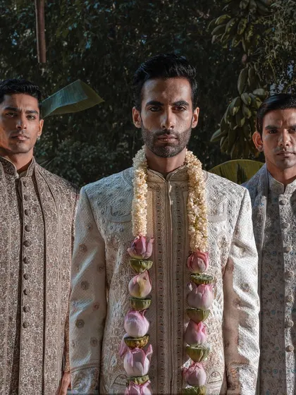 A powerful portrait of a groom flanked by his groomsmen. The focus is on the groom's majestic presence, enhanced by his finely detailed sherwani and a traditional lotus garland.