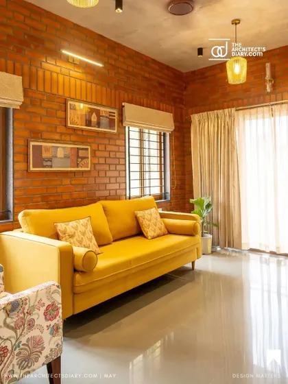 The Akshaynagar living room as featured in a publication, highlighting the vibrant yellow couch and the warm texture of the brick wall.