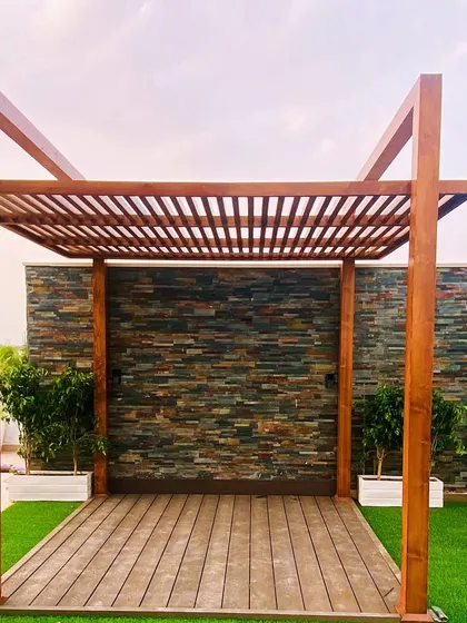 A simple wooden pergola on an artificial lawn, with a stone-clad wall providing a textured backdrop.