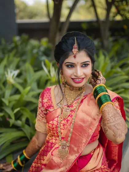 A smiling portrait of the bride sitting outdoors. The natural light brings out the rich colors of her red and green saree.