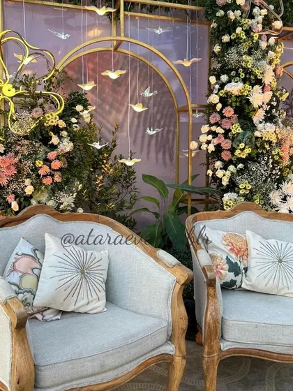 A close-up of the elegant seating at a floral-themed baby shower. The vintage-style chairs are adorned with beautiful cushions, and the backdrop features hanging birds and neon flower lights.
