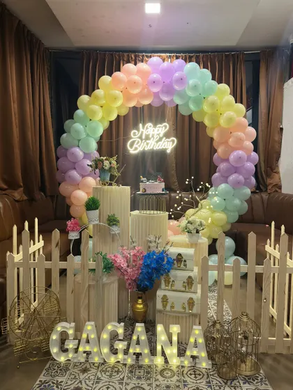A birthday party setup featuring a full ring balloon arch in pastel rainbow colors. The decor is accented with a neon "Happy Birthday" sign, a small picket fence, and marquee letters spelling the name in front.