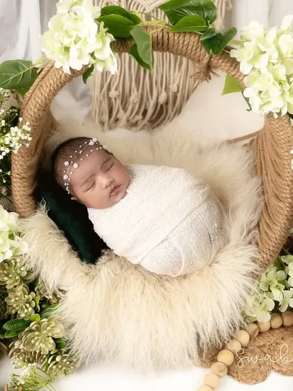 This baby is swaddled in white and nestled in a jute-wrapped basket, surrounded by white hydrangeas and pinecones. The macrame in the background enhances the bohemian theme.