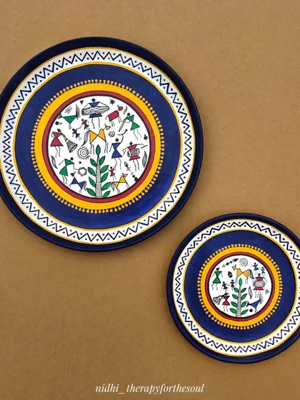 A set of two hand-painted wall plates featuring traditional Warli art. The detailed tribal figures tell a story of community and celebration, set against a deep blue background.