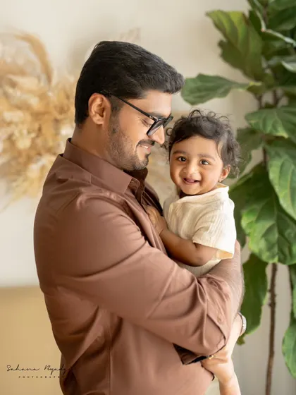 A father holds his smiling son, both looking happy and relaxed. The natural studio setting with plants adds a fresh and modern feel to this portrait.
