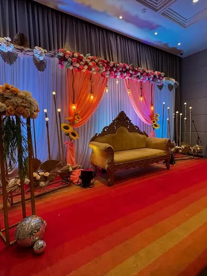 I design elegant and romantic settings for anniversaries. This stage setup features a beautiful floral arch, warm hanging lights, a classic sofa for photos, and a colorful striped carpet, creating a perfect celebratory atmosphere.